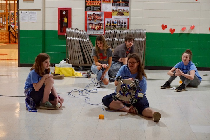 <p>Roaring Robotics adviser Tressa Kett showing the wheel structure of the mini robot. (Her son, Koby, in the background, is also a member of Roaring Robotics, Team 2704.)</p>
